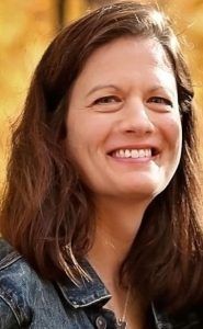 Woman with brown hair smiling, wearing a denim jacket, set against a blurred background of autumn foliage.