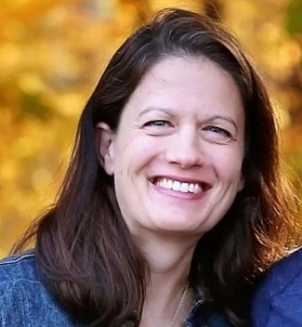 Woman with dark hair smiles, against a blurred, golden-toned outdoor background, wearing a denim jacket.