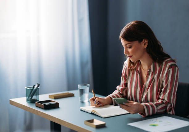 Woman writing in a notebook, holding phone, seated at a desk near a window.