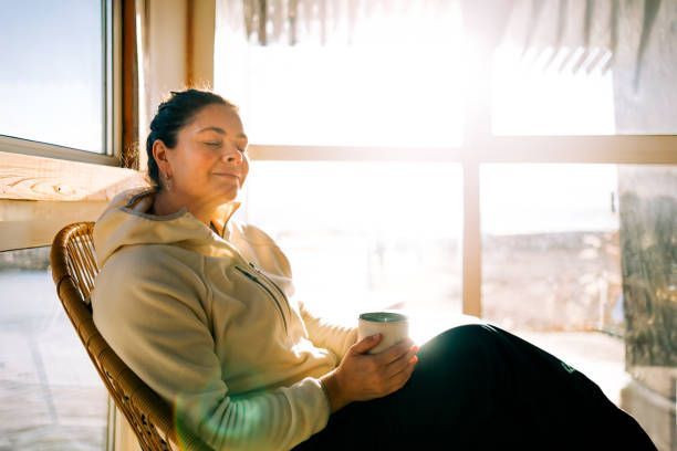Person relaxing in a sunlit room by a window, holding a mug and looking out thoughtfully