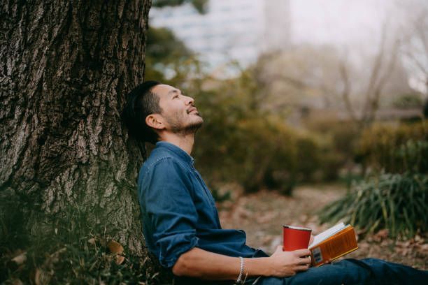 Person leaning against a tree outdoors, holding a red cup and an open book, looking up thoughtfully.