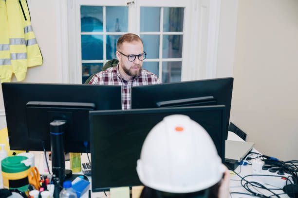 Person working at a desk with dual monitors in an office, seen behind a white hard hat in the foreground