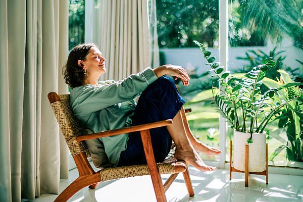 Person relaxing in a chair by a sunny window with green houseplants and curtains