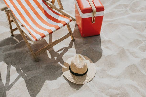 Striped beach chair, red cooler, and straw hat on sandy beach sand.