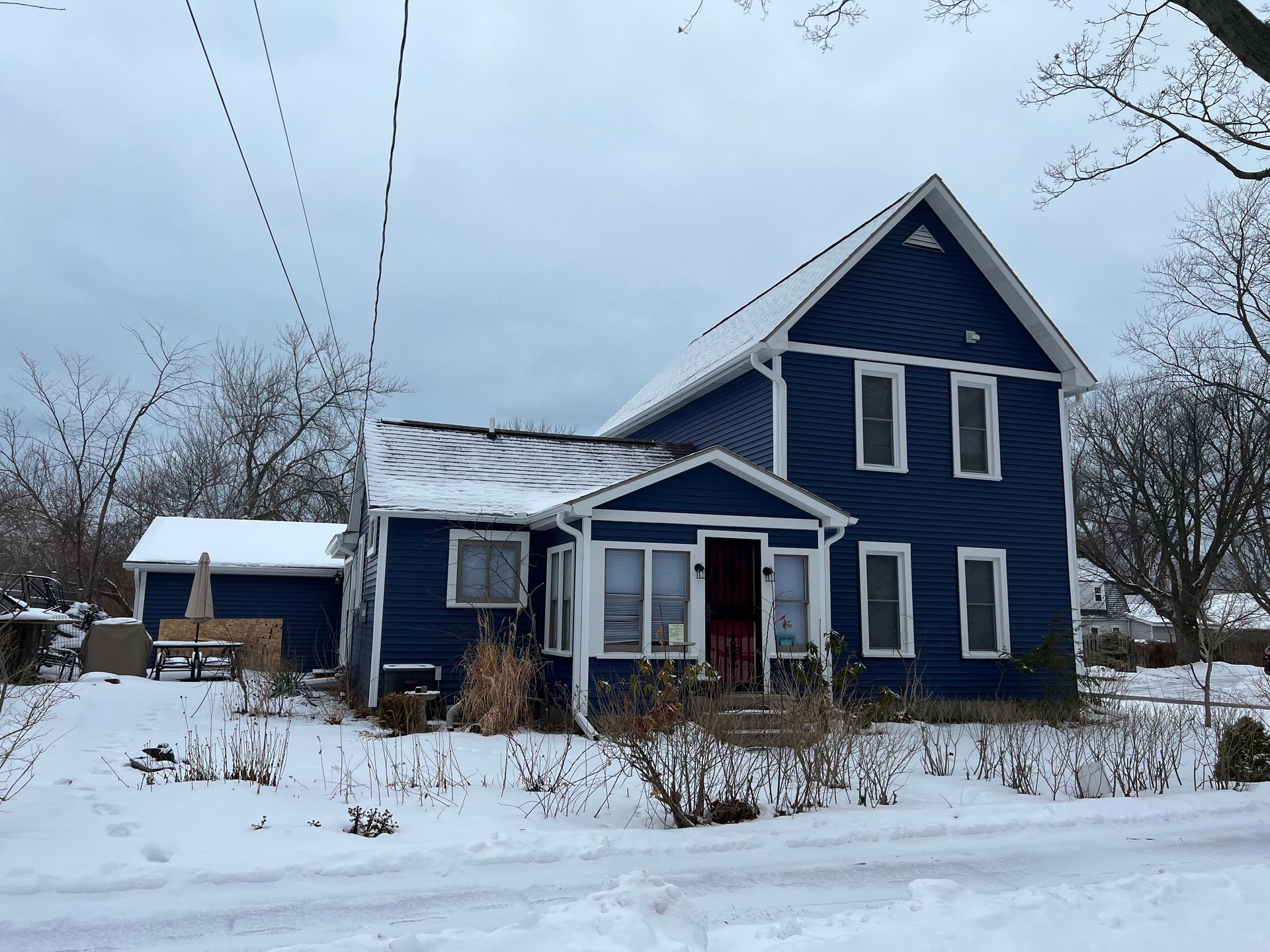Blue house with snow-covered roof and yard in winter, with bare trees and power lines overhead