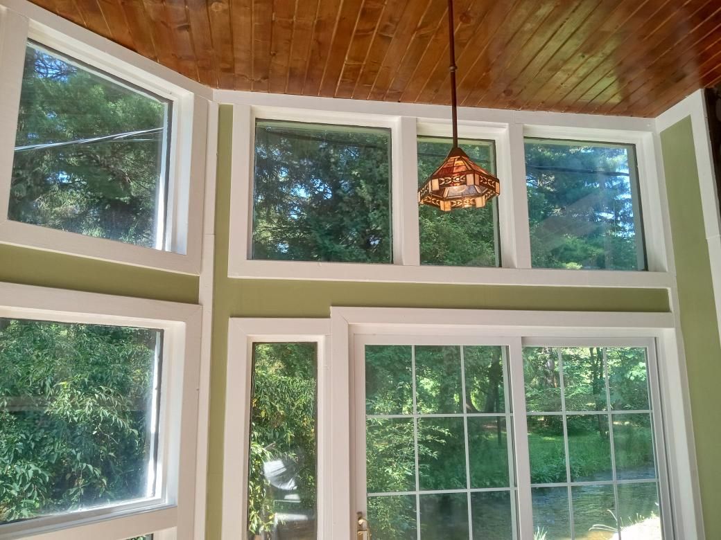 Sunlit room with large windows, white trim, green forest view, and a wooden ceiling with pendant light