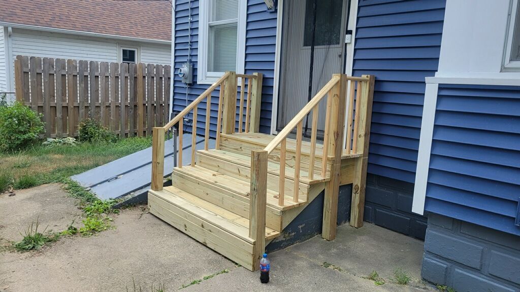 Small wooden accessibility ramp and steps attached to a blue house beside a concrete driveway.