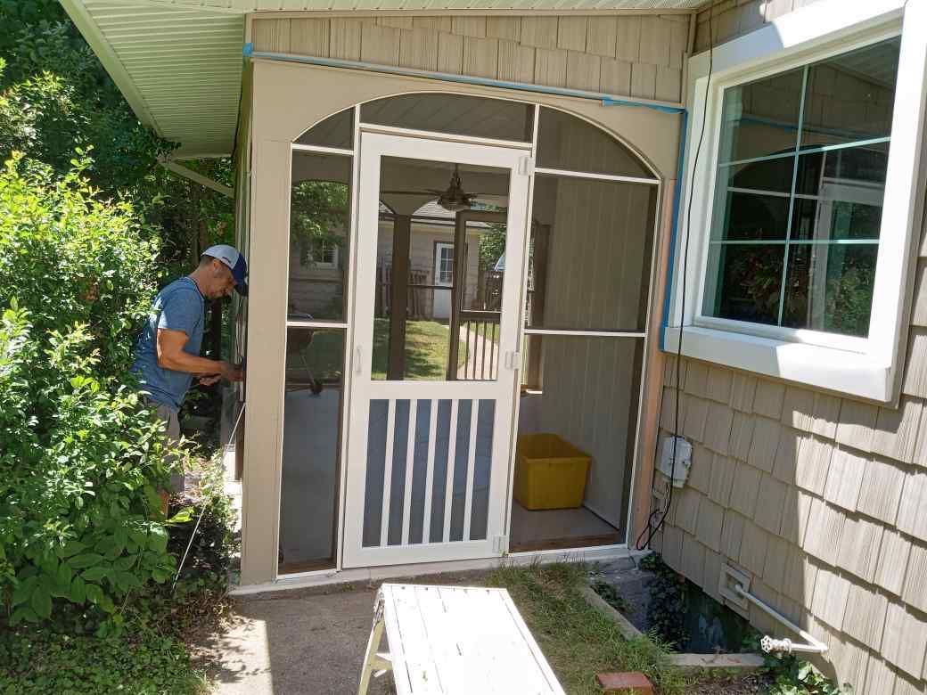 Person standing by a screened front door on a house porch, with bushes and a window nearby.