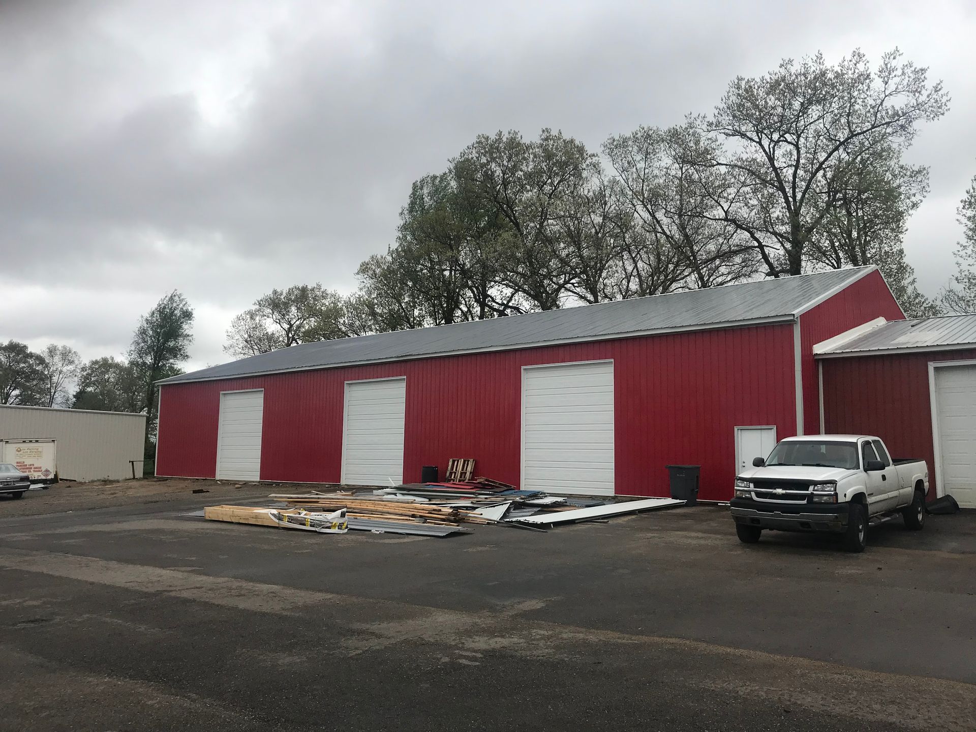 Red warehouse with white garage doors and a white pickup truck in a parking lot under cloudy skies