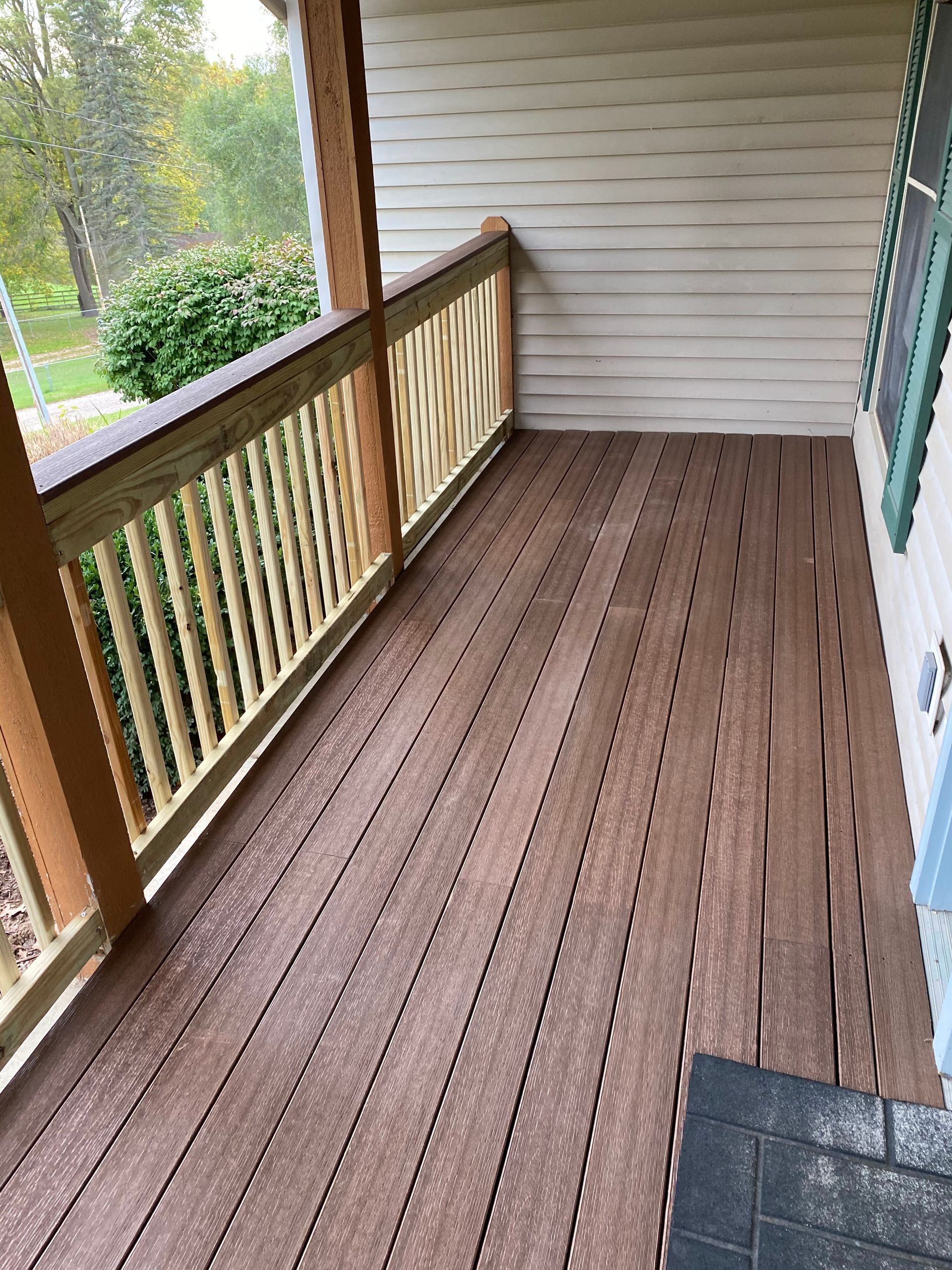 Empty wooden porch with railing, brown floorboards, and white siding overlooking trees.