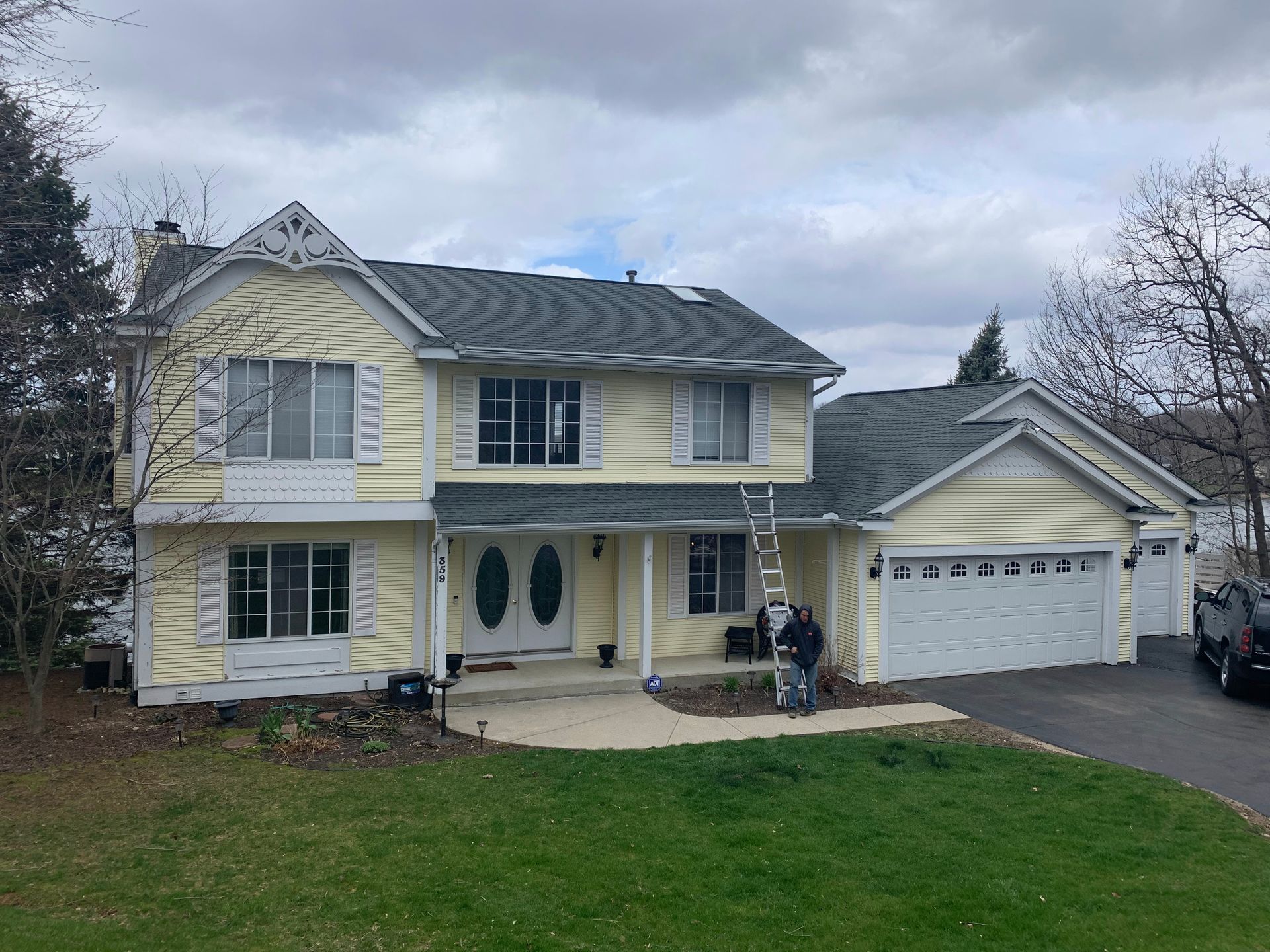 Two-story yellow house with white trim, attached garage, and a green lawn under a cloudy sky