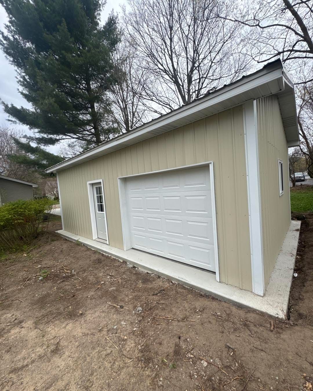 Small beige garage with white door beside a dirt driveway, viewed from the front under cloudy skies
