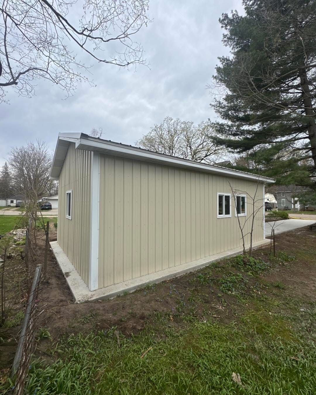 Beige shed with white trim in a grassy yard, viewed from the side under cloudy skies