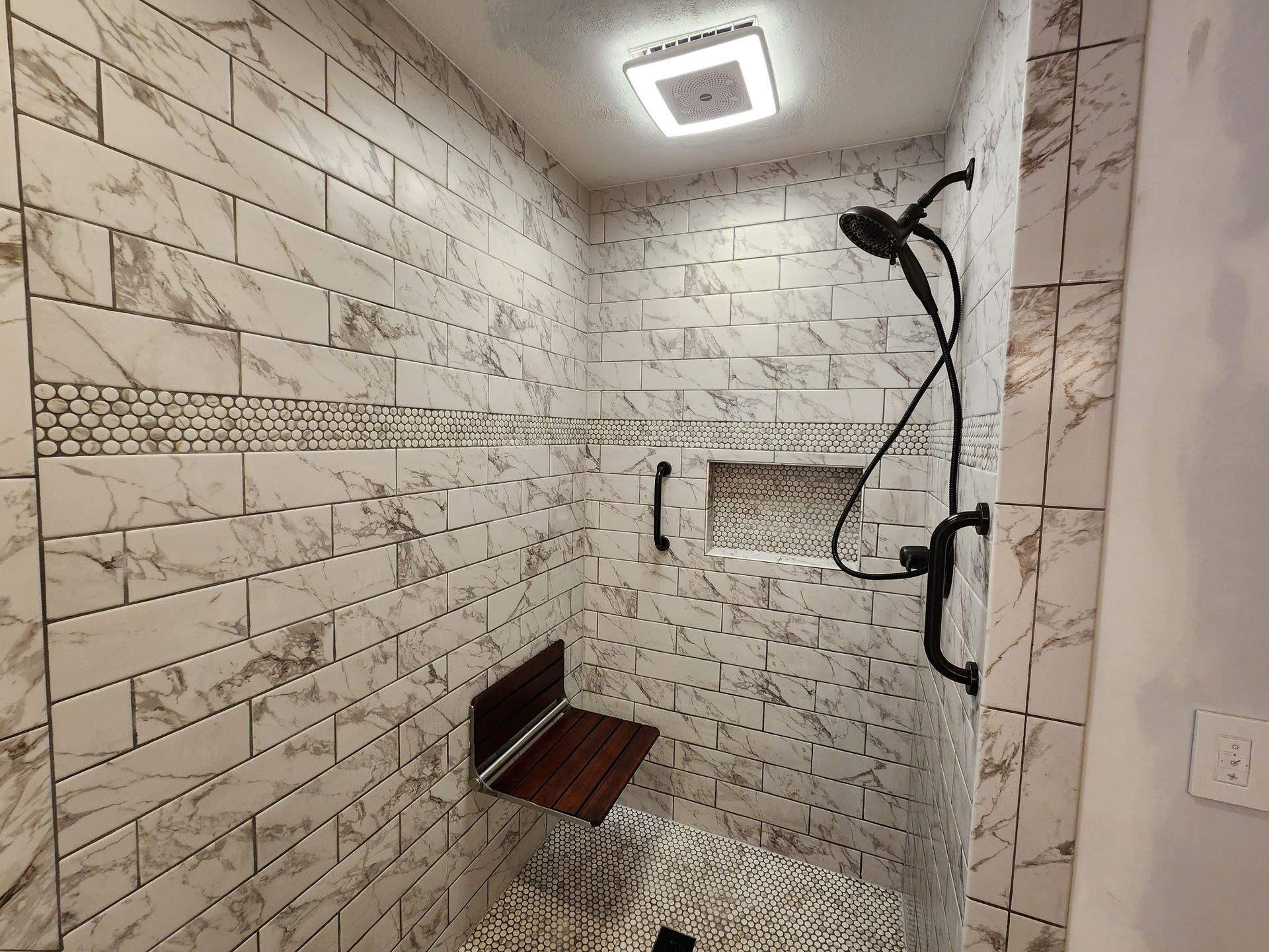 Modern tiled shower with bench, glass shelf, and black shower fixtures under a square ceiling light