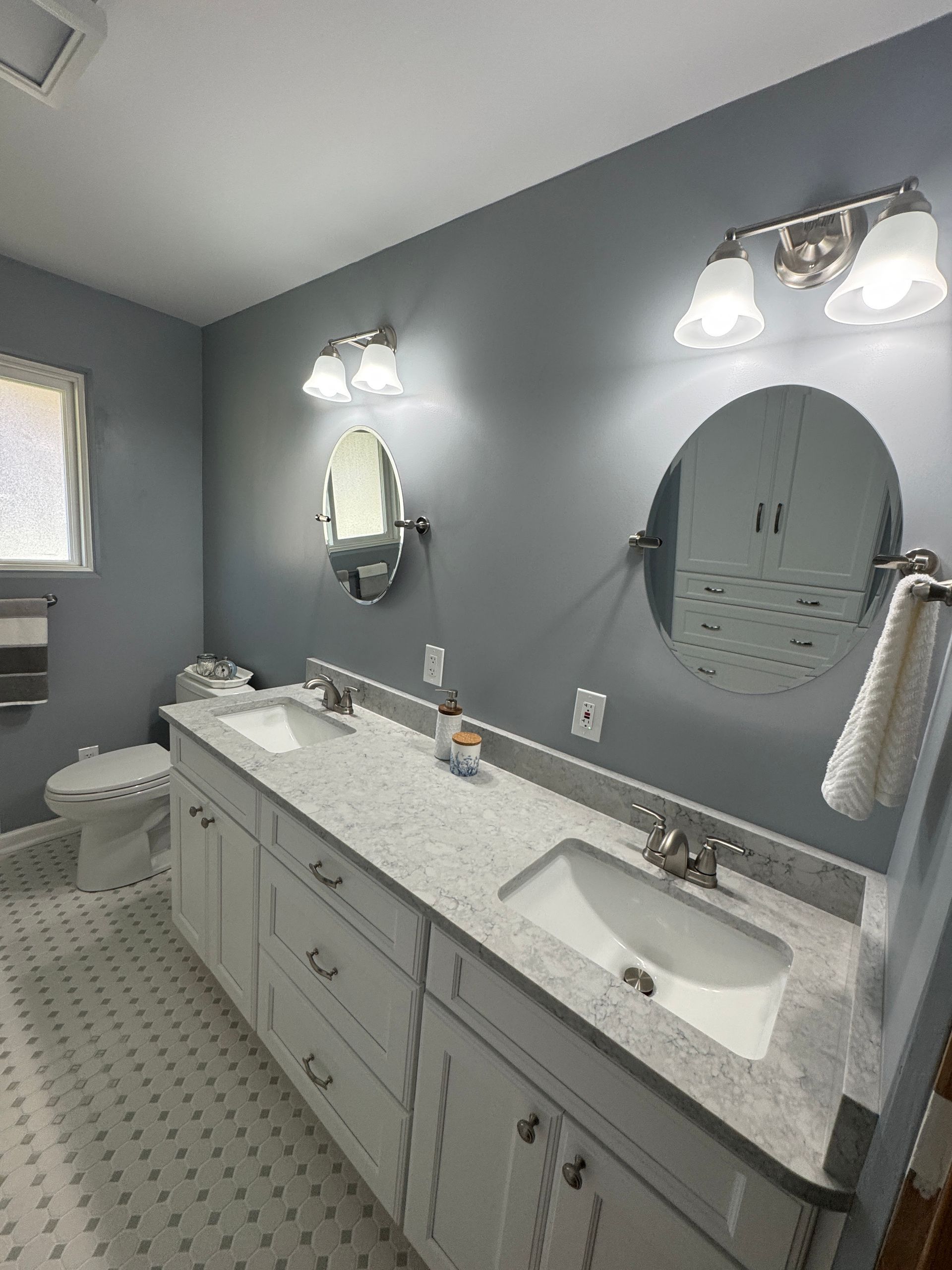 Gray bathroom with double vanity, round mirror, and wall sconces, shown from the doorway.