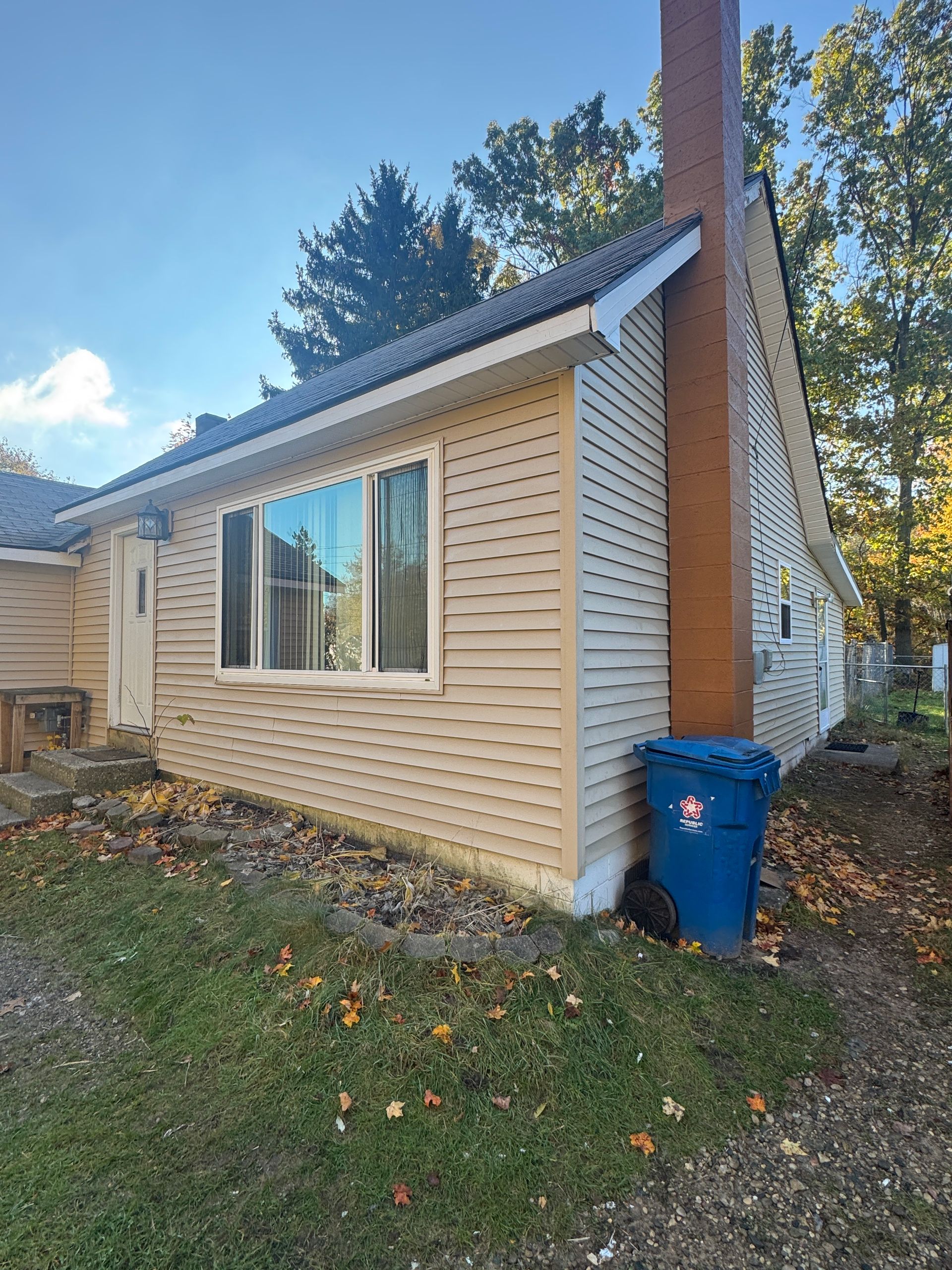 Tan beige house exterior with a large window, chimney, and blue recycling bin beside the wall.