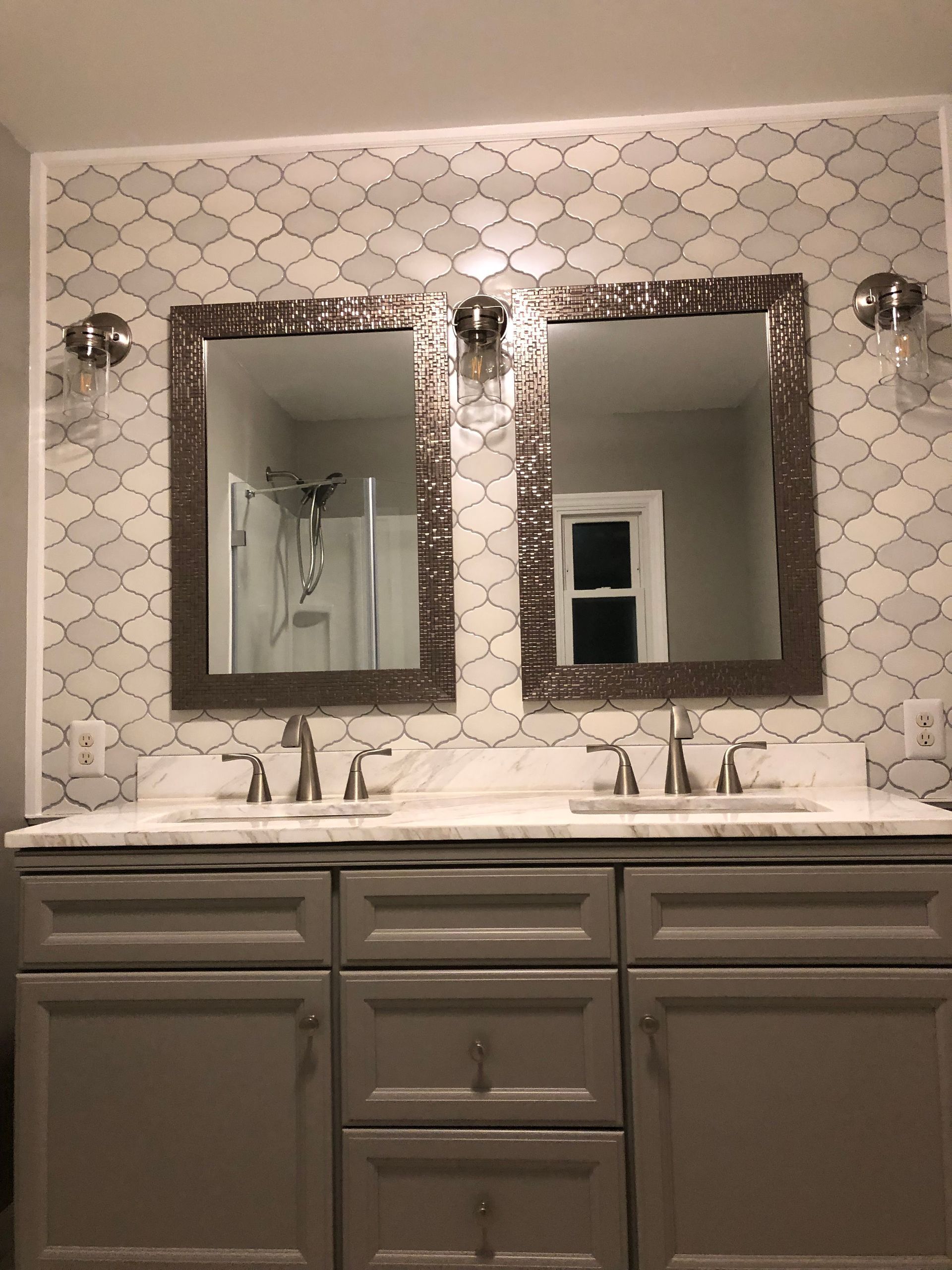 Bathroom vanity with double mirrors, chrome sconces, and gray cabinets against patterned tile wall