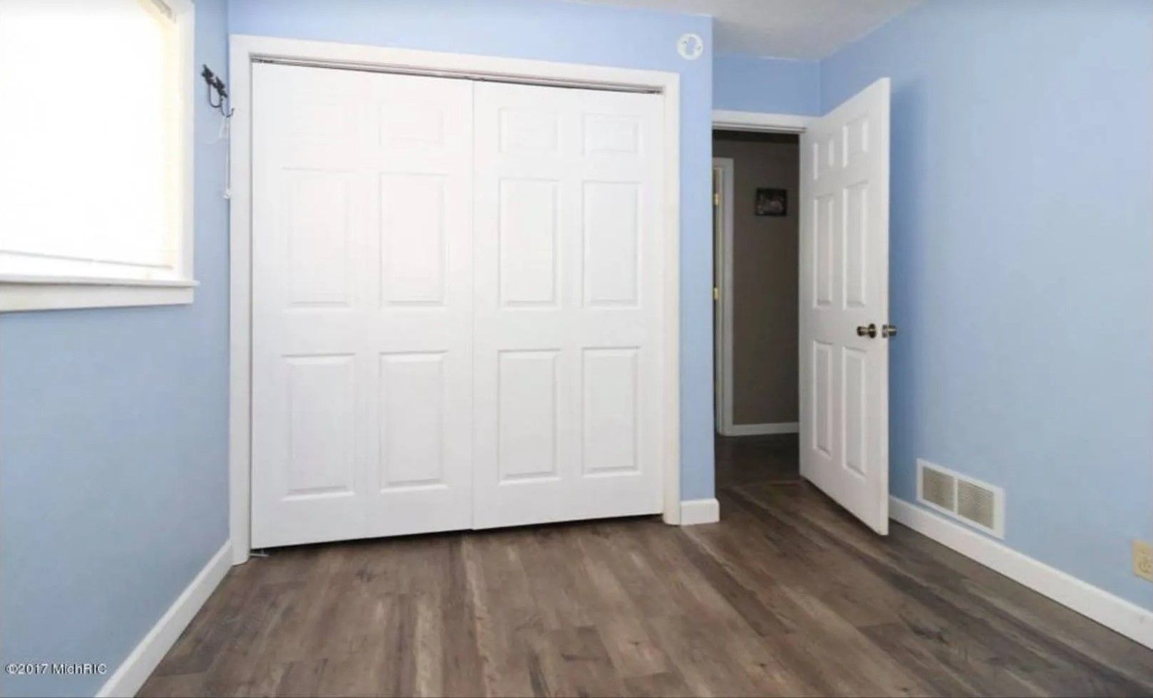Empty bedroom with light blue walls, wood floor, and white closet doors.