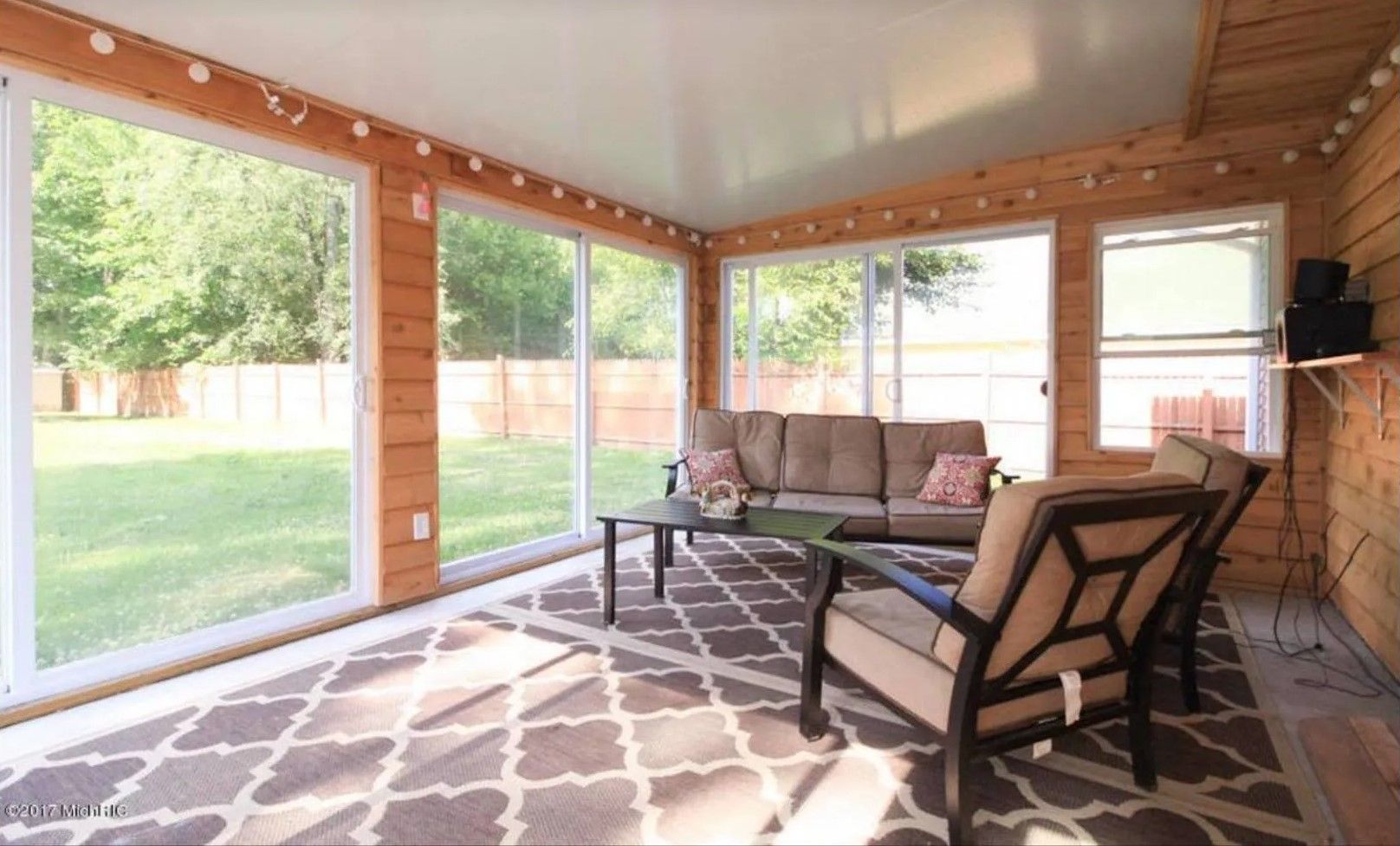 Sunroom with large windows, wicker sofa, lounge chair, and patterned rug on a wooden floor.