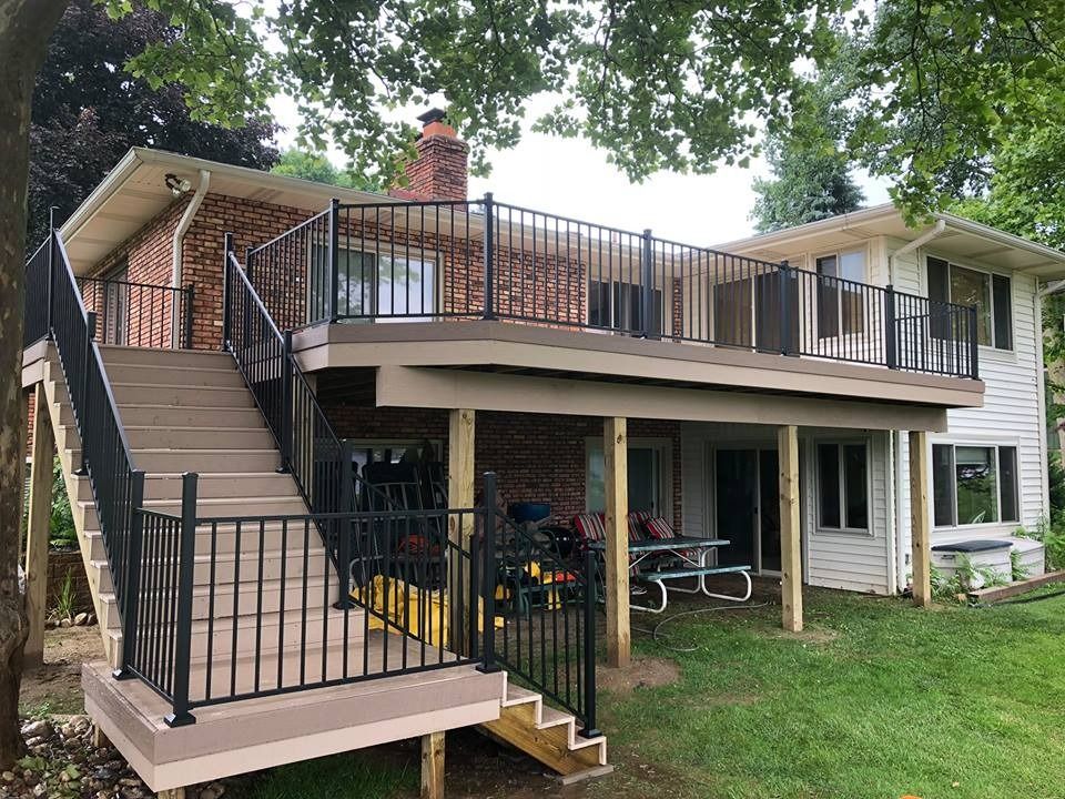 Two-story house with a large wraparound deck, exterior staircase, and black railing under trees.