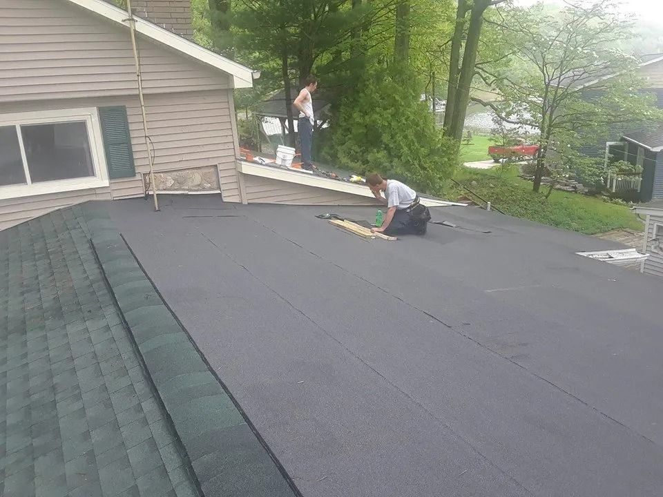 Roofing workers installing a dark shingle roof beside a house with trees in the background