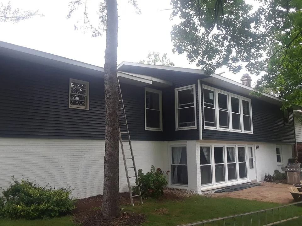 Two-story house with dark siding, white lower level, and a ladder leaning against the front tree.