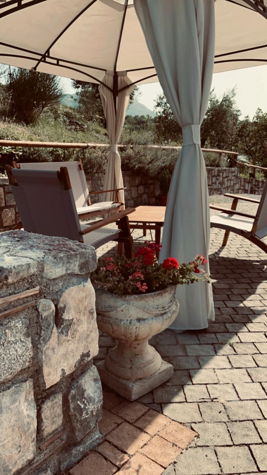 Tavolini da patio sotto un ombrellone color crema accanto a muri in pietra e una fioriera al sole