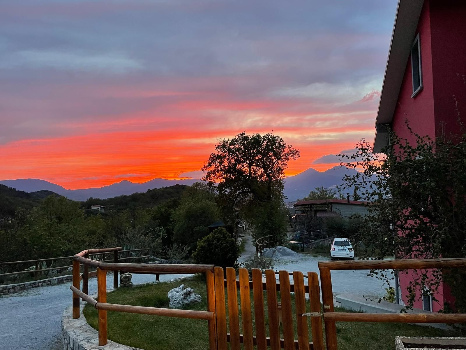 Tramonto sulle montagne con un cancello di legno, un sentiero di ghiaia, alberi e una casa rosa sulla destra.
