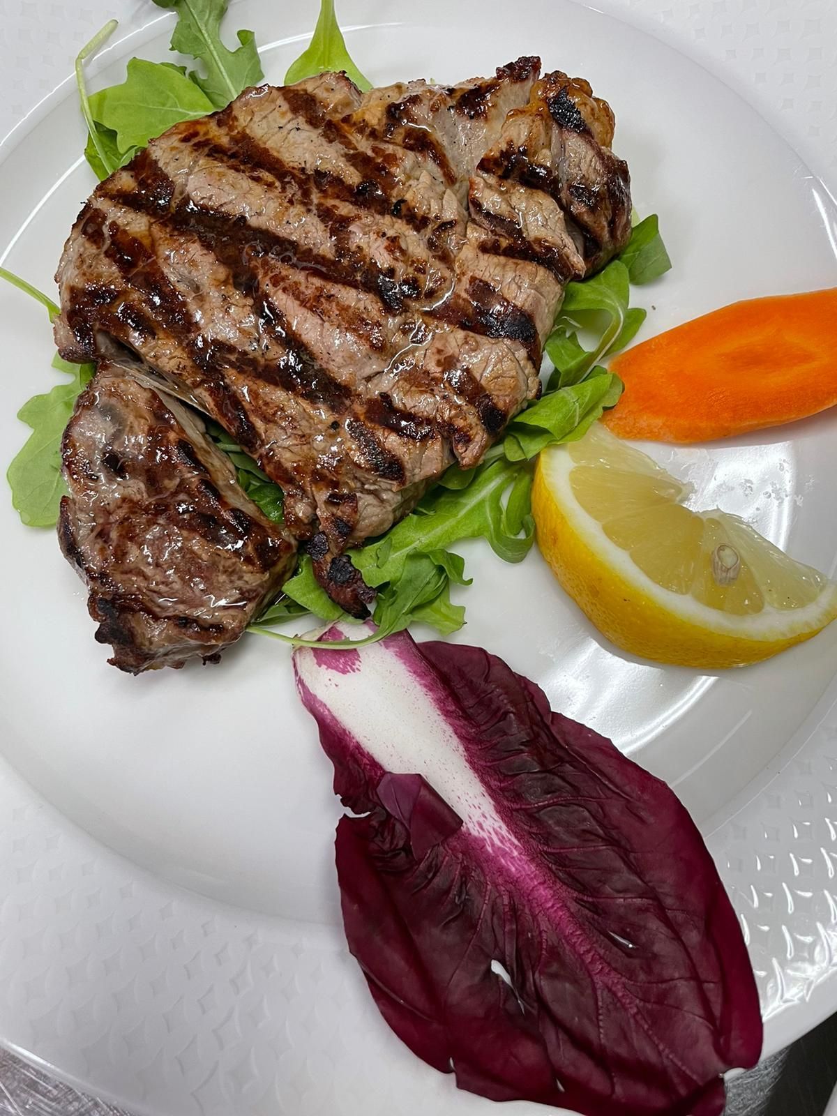 Bistecca alla griglia su un piatto bianco con verdure, spicchio di limone, fetta d'arancia e radicchio