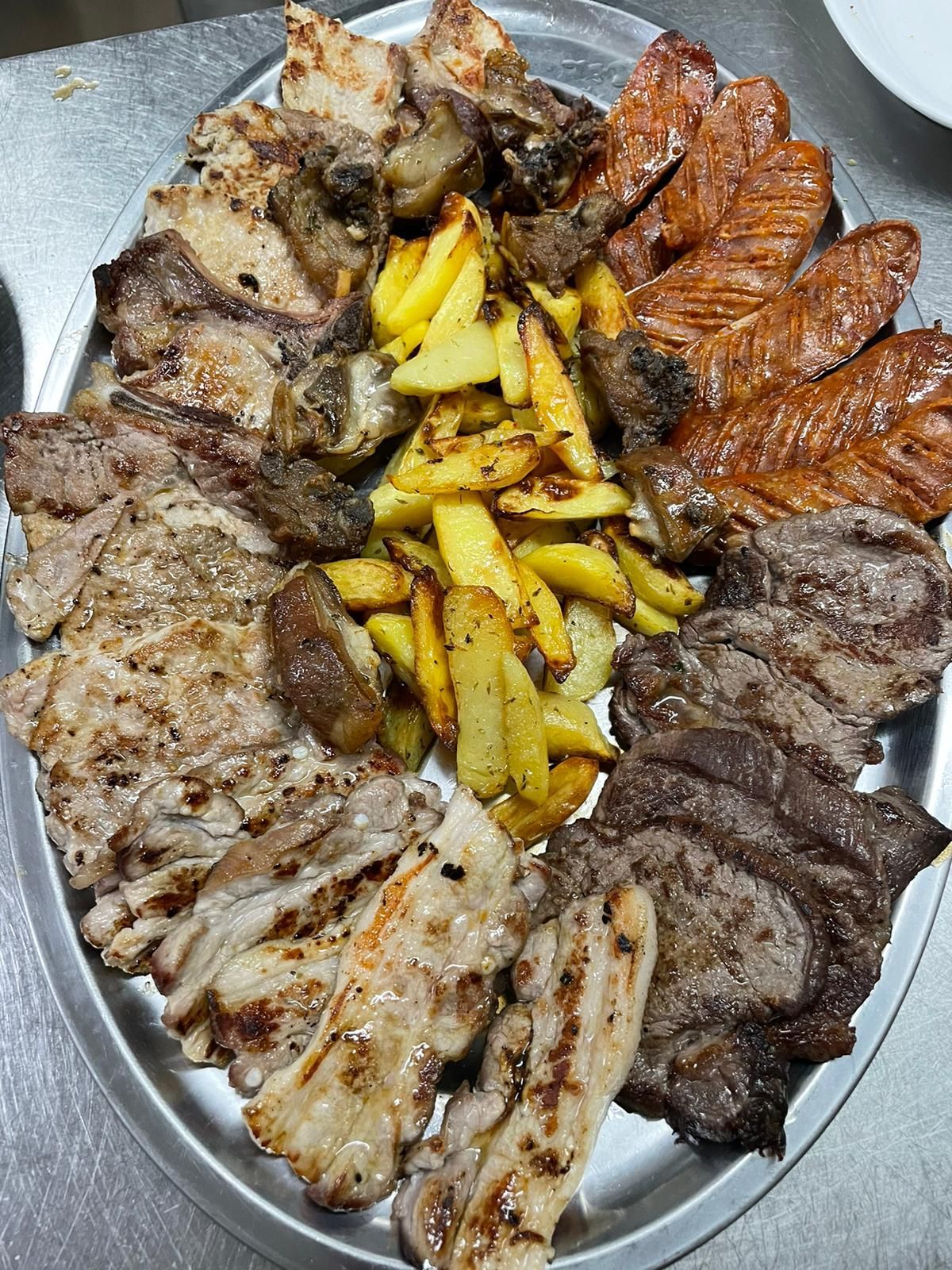 Piatto con un assortimento di carni grigliate e patatine fritte, disposto su un vassoio ovale di metallo.