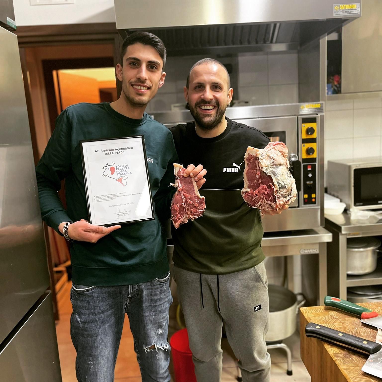 Due uomini in cucina, sorridenti accanto ai fornelli, tengono in mano della carne cruda e un certificato.
