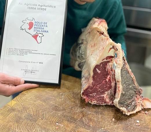 Un certificato incorniciato è appoggiato su un tavolo accanto a un grosso taglio di carne, tenuto in mano da una persona con una camicia verde.