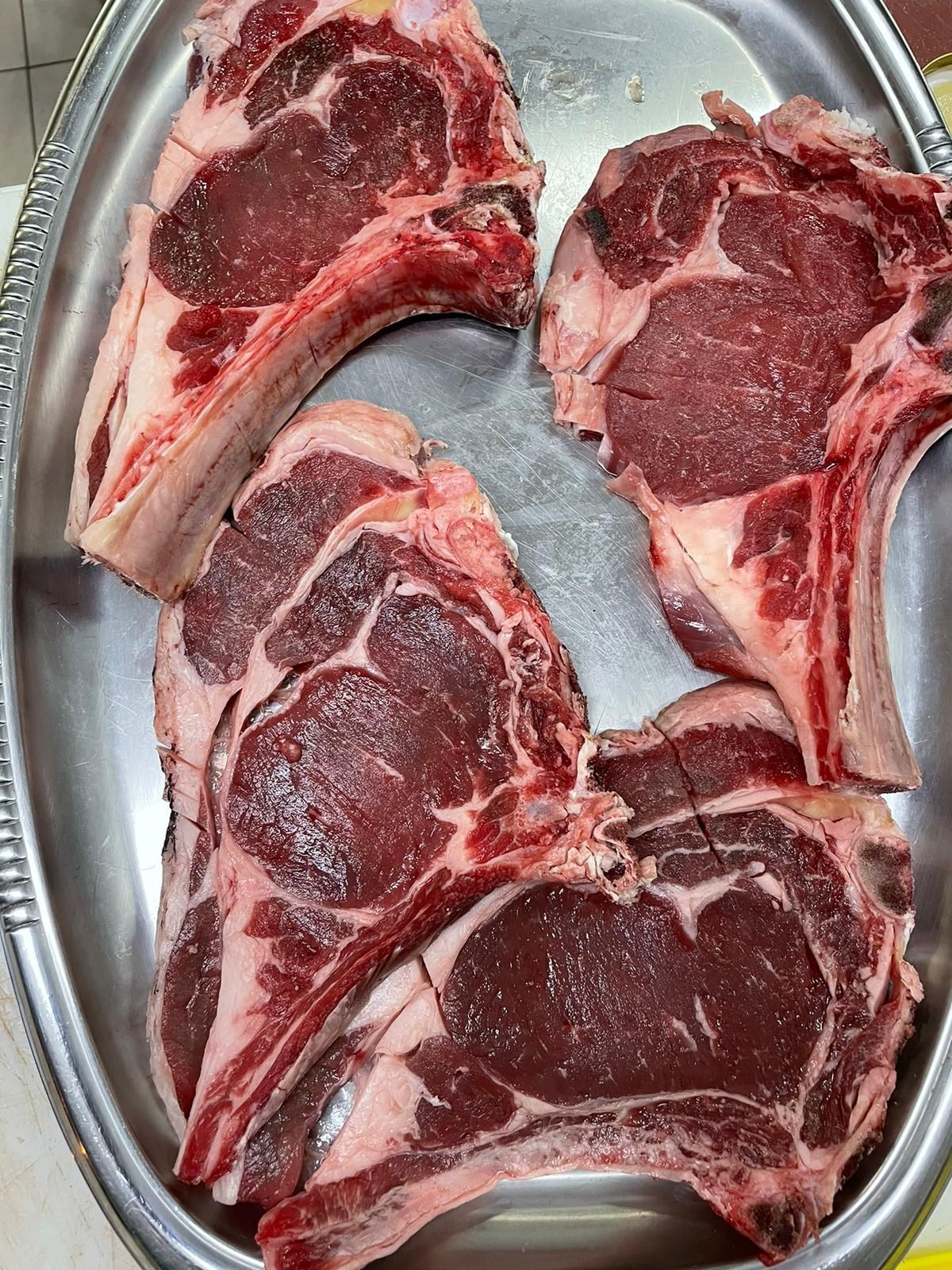 Quattro costolette di agnello crude su un vassoio d'argento, carne di un rosso intenso con grasso bianco e ossa.