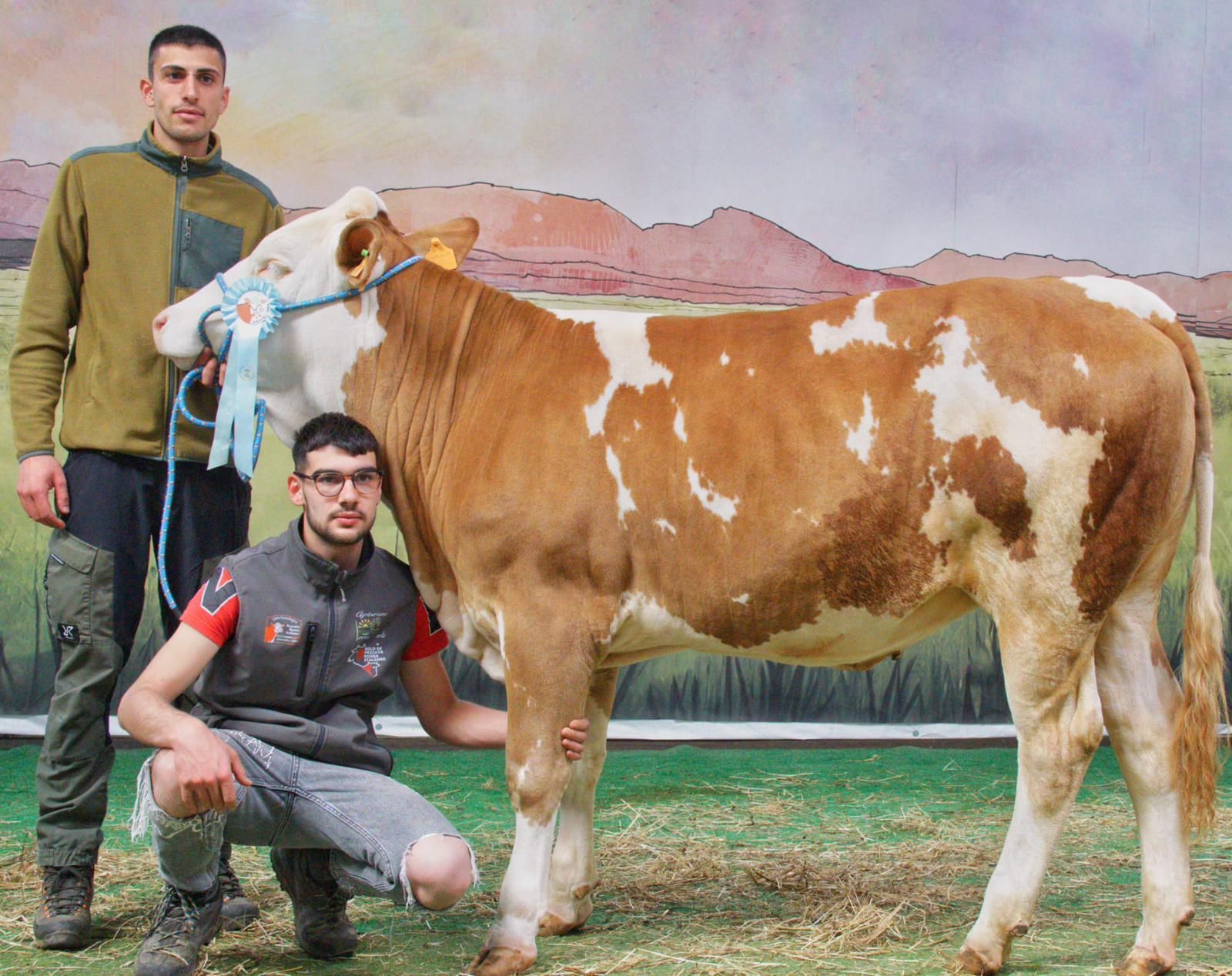 Due uomini posano con una mucca marrone e bianca in un prato all'aperto, con le montagne sullo sfondo.