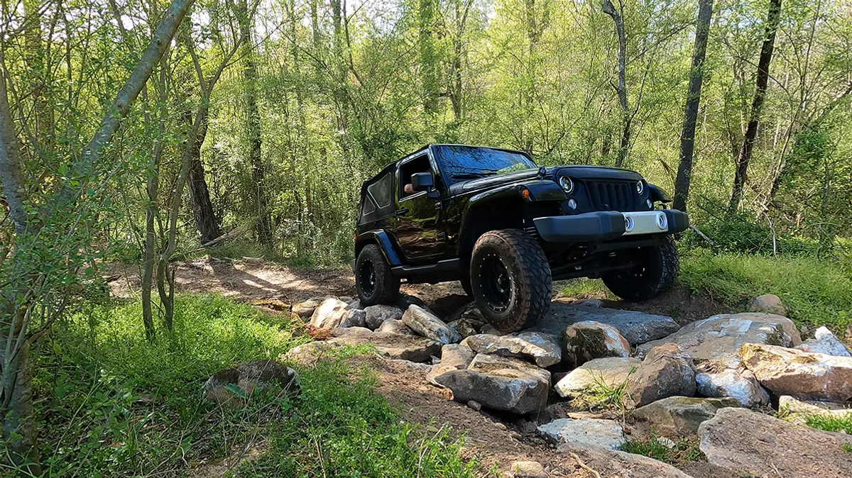 A black jeep is driving down a rocky trail in the woods.
