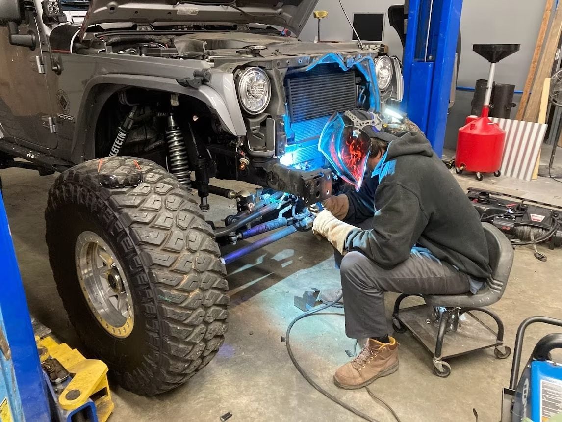 A person wearing a welding helmet works on the suspension of a lifted gray Jeep inside a professional automotive shop.