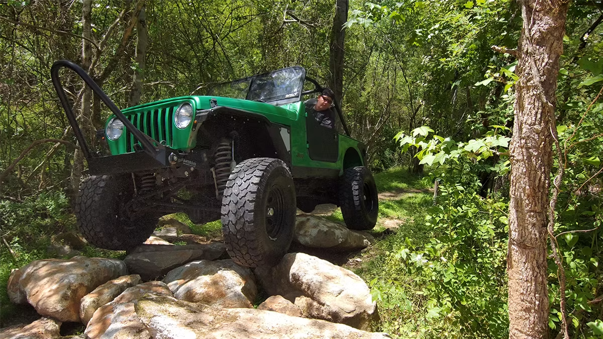 A green jeep is driving down a rocky trail in the woods.