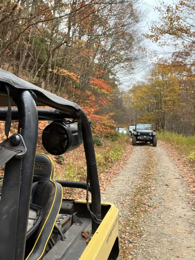 Yellow off-road vehicle leads a line of vehicles on a dirt road in a fall forest setting.