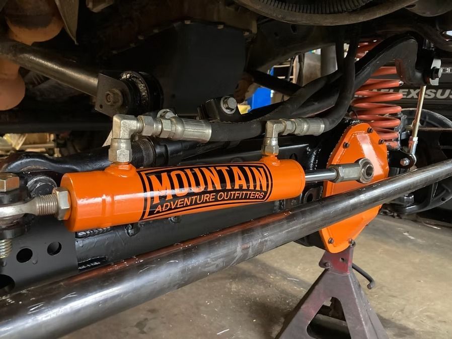 Bright orange hydraulic steering ram mounted on a vehicle's front suspension axle, labeled