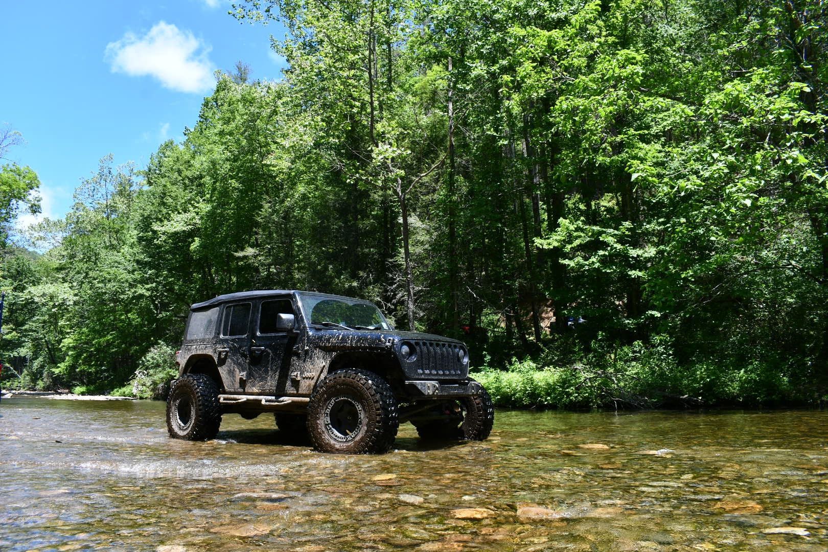 A muddy black Jeep driving through a shallow, rocky river surrounded by a lush green forest under a blue sky.