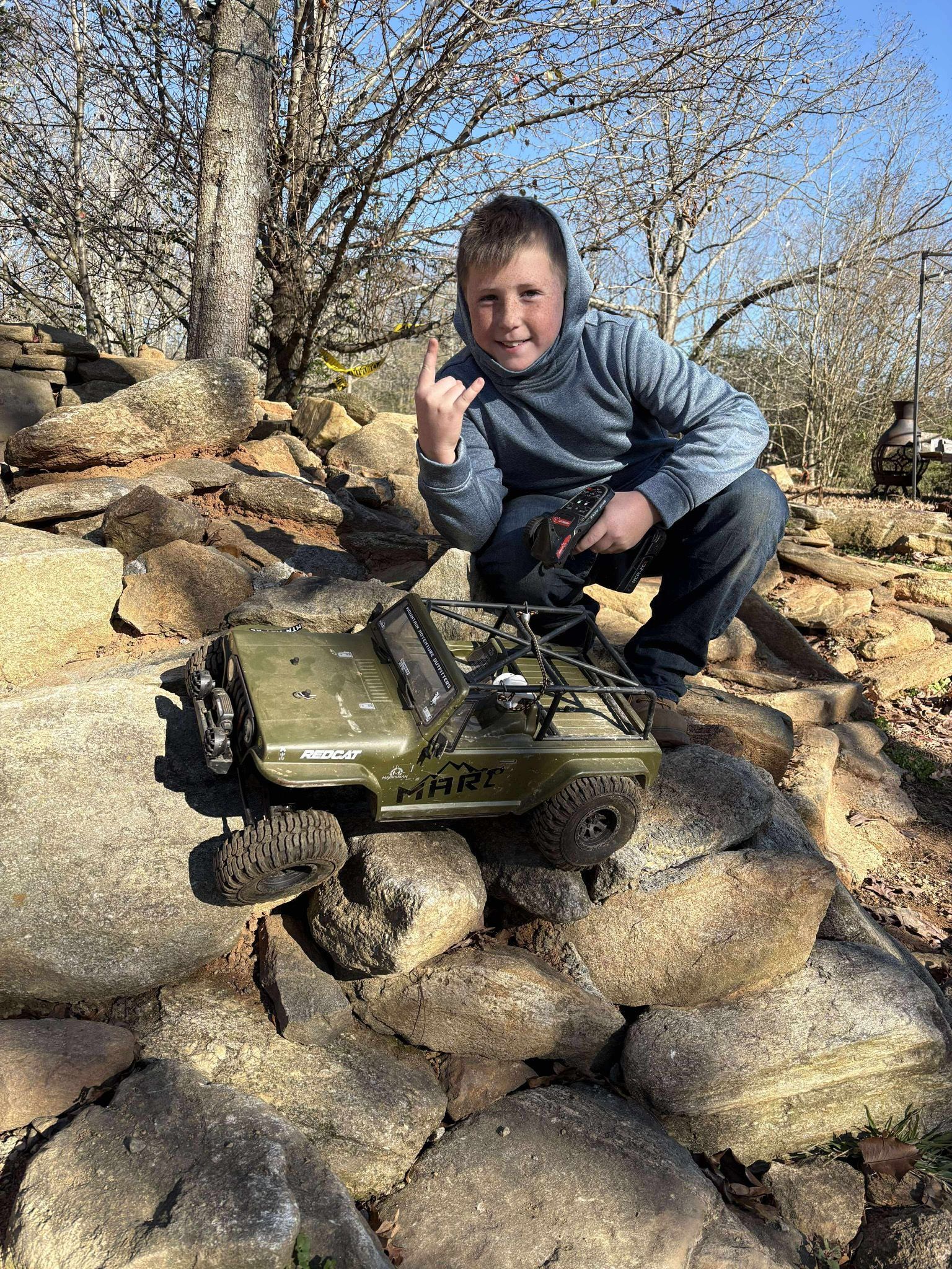 Boy crouching on rocks beside a small green remote-controlled tank outdoors on a sunny day