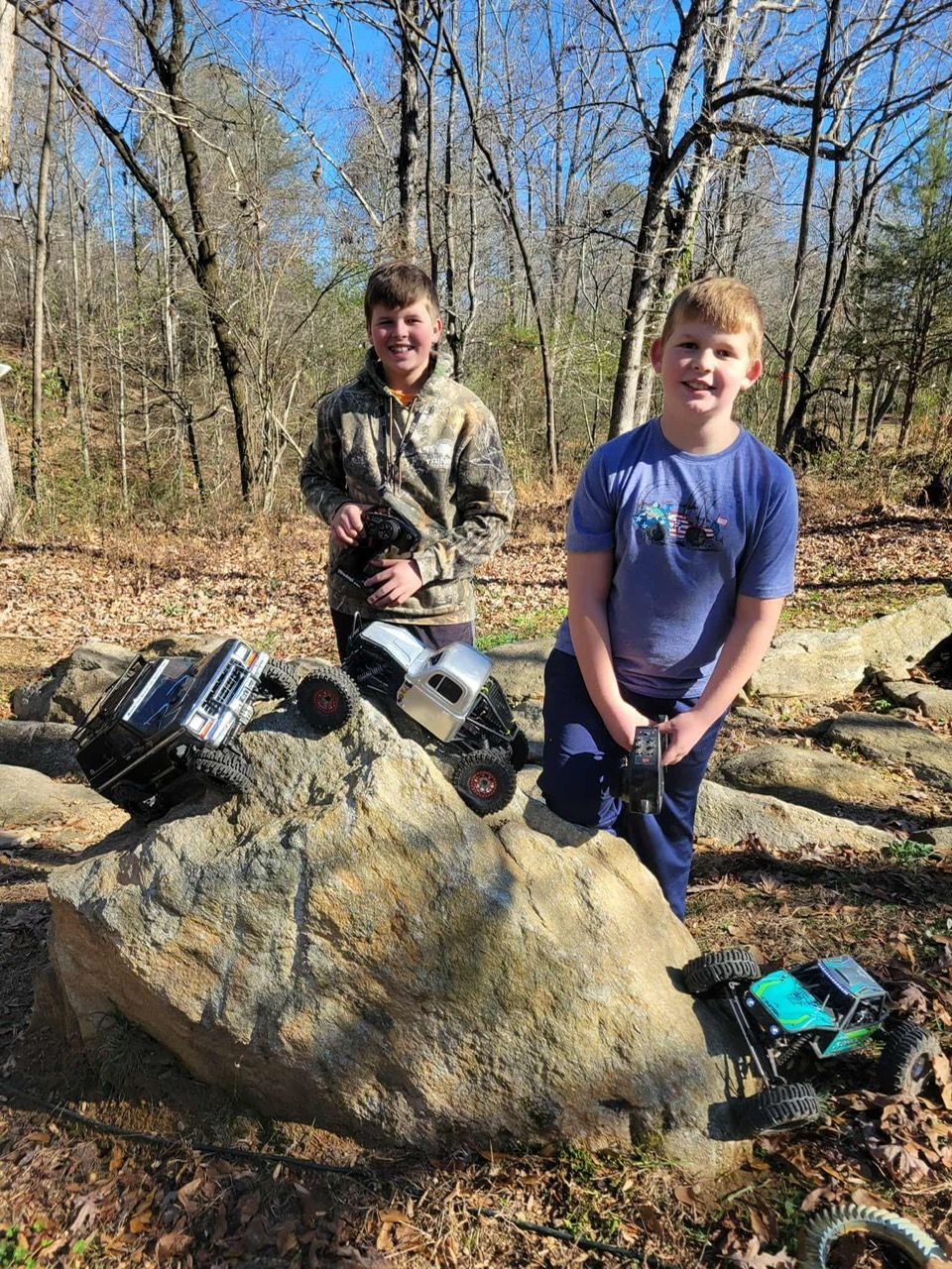 Two people standing by a large rock with remote-control cars in a wooded area.