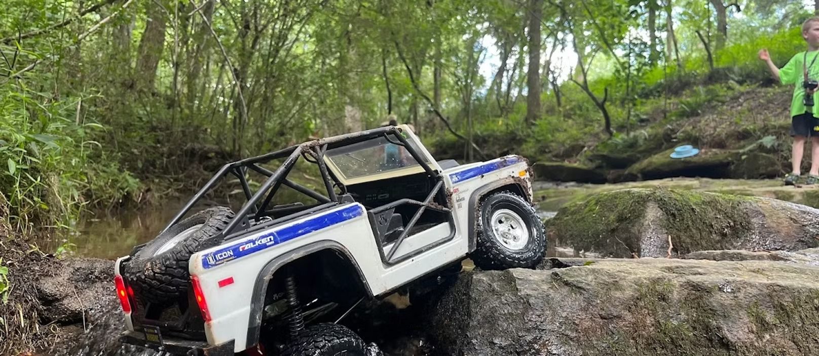 White off-road jeep stuck on rocks in a muddy forest creek, with a person standing nearby