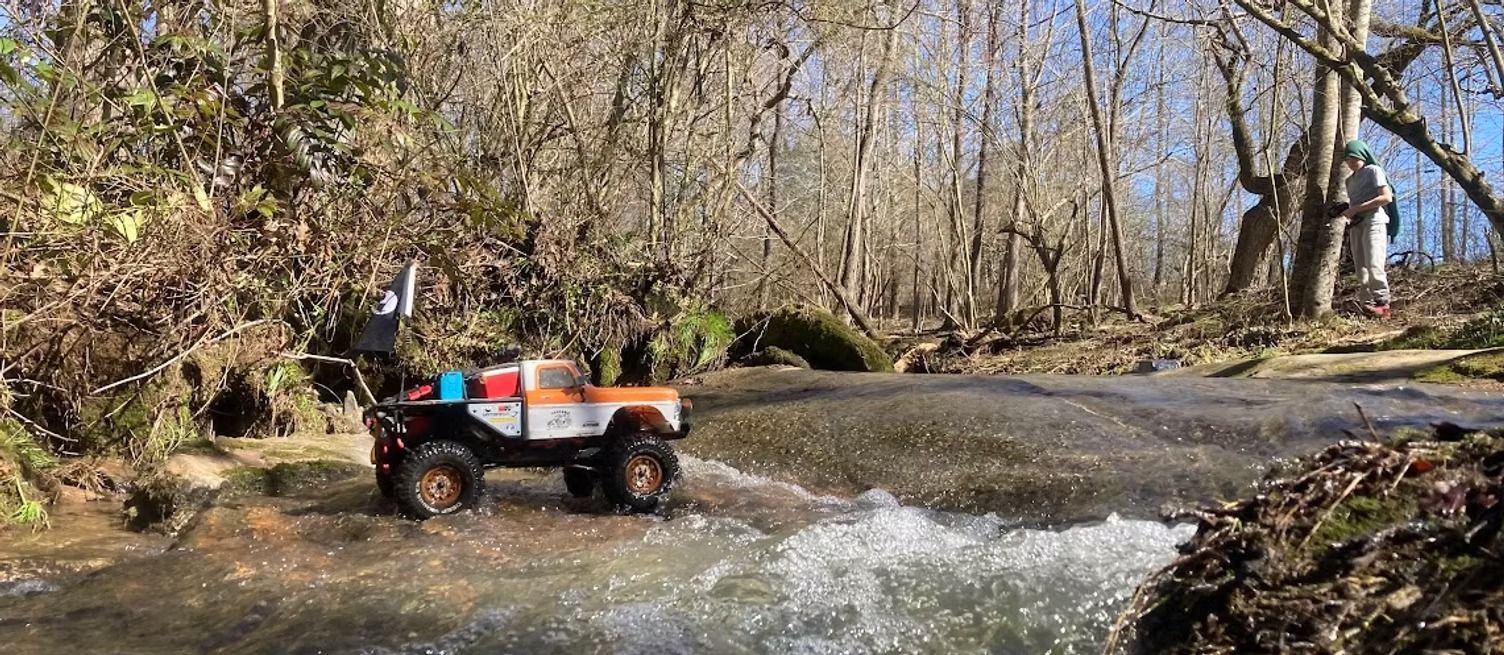 Orange off-road truck crossing a shallow creek in a wooded area