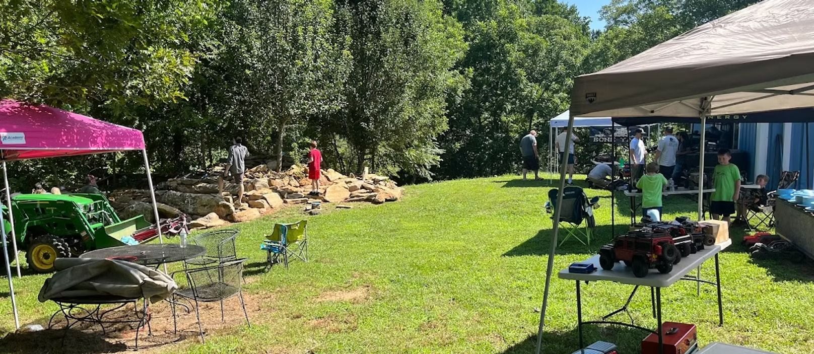 Outdoor backyard gathering with tents, tables, and people grilling on a sunny lawn.