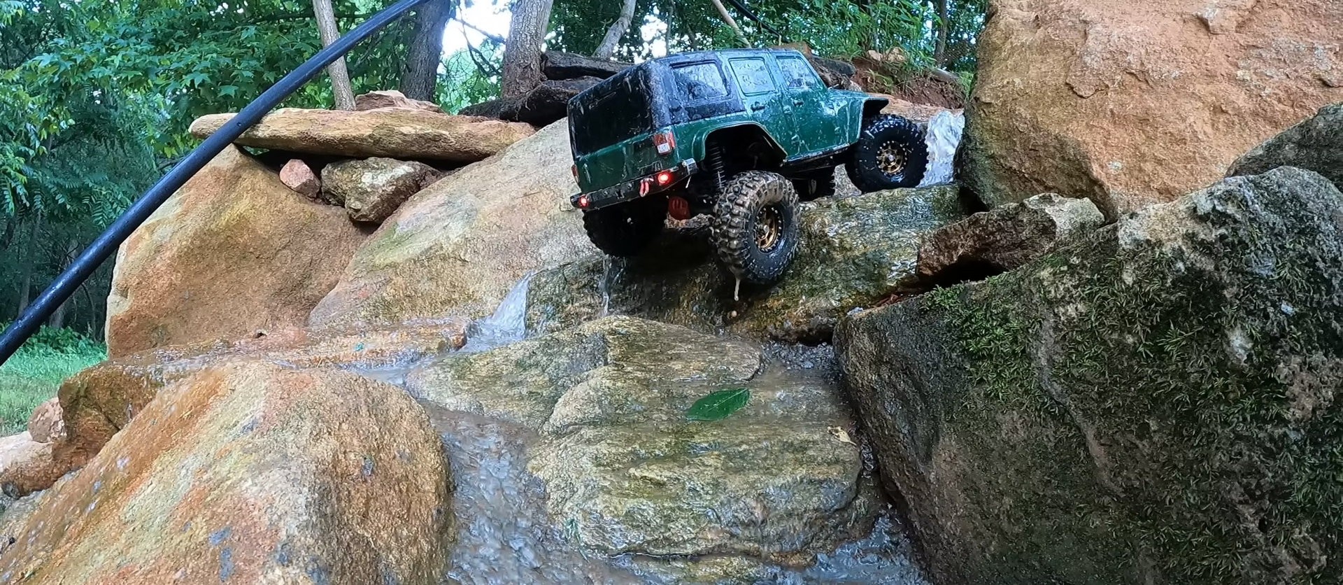 Blue toy off-road truck climbing wet rocks under a garden hose stream