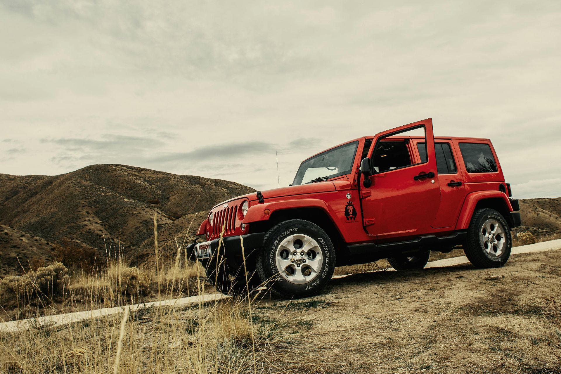 A red Jeep Wrangler sits parked on a dry, grassy hillside under a cloudy sky.