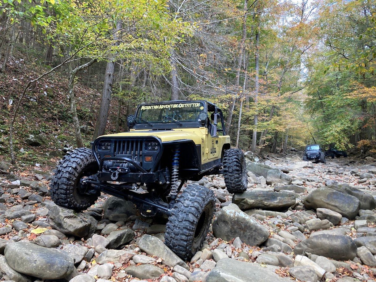 Tan Jeep rock crawling through a rocky, wooded stream.