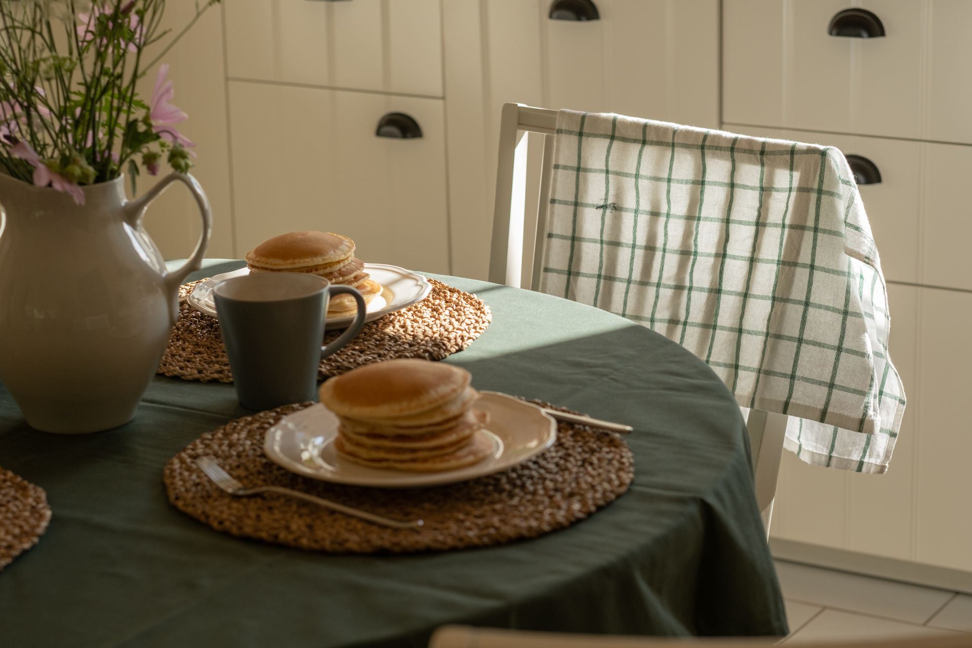 A table set with pancakes, mug, vase of flowers, and a checkered towel draped on a chair.