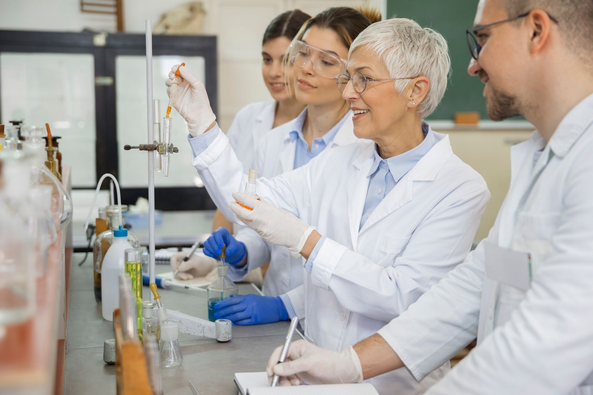 Laboratory group examining a test tube. Scientists wear white lab coats, gloves, and safety glasses.
